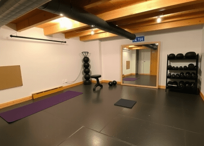 Basement gym room with rubber flooring and exposed wooden beams, featuring neatly arranged exercise equipment, a large mirror, a yoga mat in one corner, and a shelf with weights, creating a rustic and encouraging workout atmosphere.